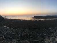 A wide-angle scene of a rocky coastline at twilight, with calm waters and a pastel gradient sky, dark rocks in the foreground, and distant headlands across the horizon.