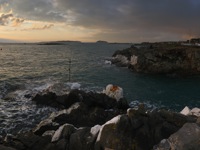 A rocky coastal inlet at sunset, with swirling ocean waves, rugged cliffs, and a small marina in the background, under a sky with dramatic cloud cover and rays of sunlight breaking through