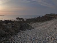 A panoramic view of a rocky and pebbly storm beach at sunset, with the sun low on the horizon over the ocean, rugged rocks in the foreground, and a distant stone building along the shoreline.