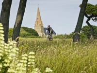 A cyclist riding along a grassy, flower-dotted path with tall trees framing the view and Vale Church spire in the background beneath a blue sky.