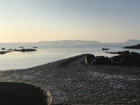 A stone slipway leading into a tranquil, early morning sea, with a few small boats anchored nearby and gentle hills silhouetted in the hazy distance.