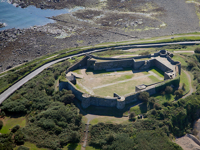 An aerial view of Vale Castle with thick defensive walls, surrounded by Castle Road coastal road, rocky shoreline, and lush vegetation.