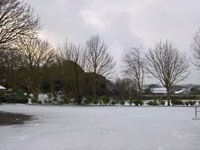 Vale Douzaine Rooms parking area after snowfall, with bare winter trees, snow-covered ground, and buildings in the background beneath a cloudy sky.