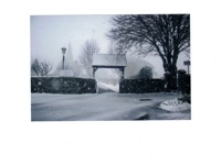 A tranquil winter scene of Vale Church entrance in snowfall, with a lychgate, bare trees, lamp post, and soft snow covering the ground and rooftops.