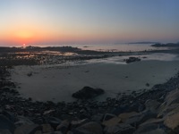 A panoramic view of a rocky, tidal shoreline at dusk, with scattered pools and wet sand reflecting the orange glow of the setting sun on the left.