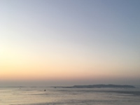 A serene coastal scene at dusk with calm waters, rocky shoreline in the foreground, distant headlands silhouetted by a fading sunset, and two jet contrails visible in the sky.