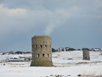 Two round stone Martello towers set in a snowy landscape at L’Ancresse, Guernsey, with houses and trees on the horizon under an overcast sky.