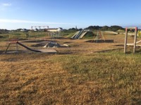 An empty playground under clear blue skies with dry grass, featuring swings, slides, climbing frames, and picnic benches, with buildings and Vale Church spire visible in the distance.